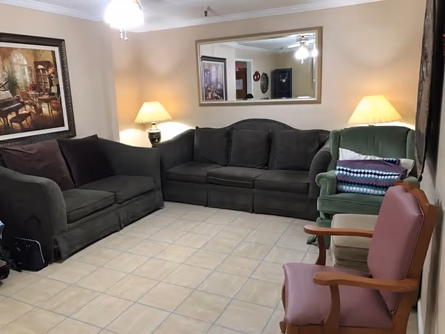 Spacious living room with tiled floor, green upholstered sofas and armchairs, side lamps and a large wall mirror.