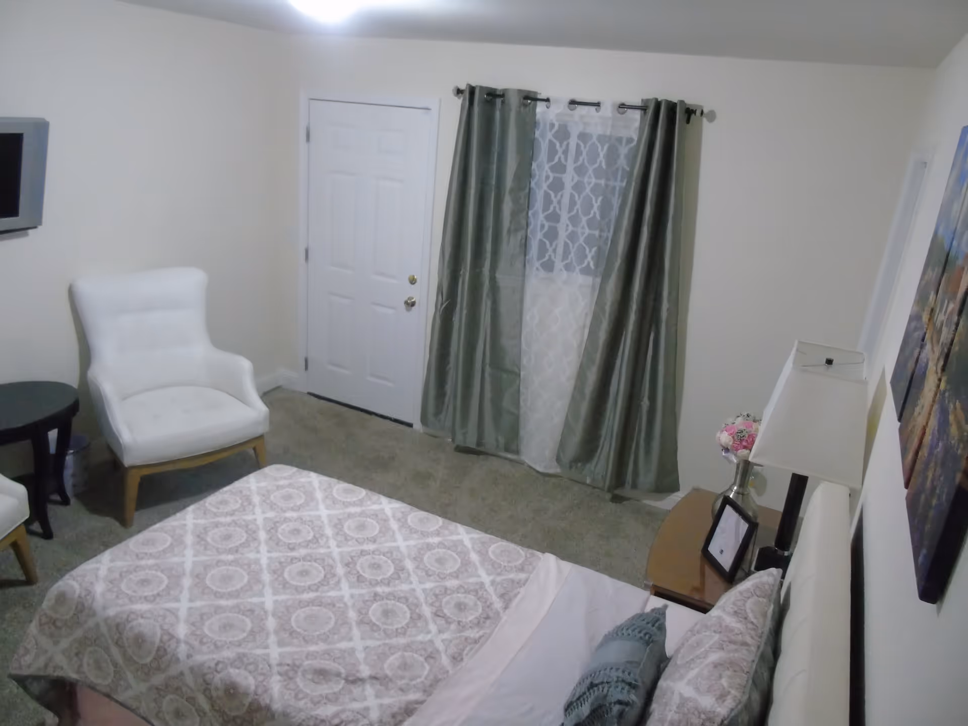 A bedroom with a bed covered in a patterned bedspread and pillows, a white upholstered chair next to a small round table, a window with sheer and green curtains, a white door, a bedside table with a lamp, a vase of flowers, and a framed photo, and a wall-mounted TV.