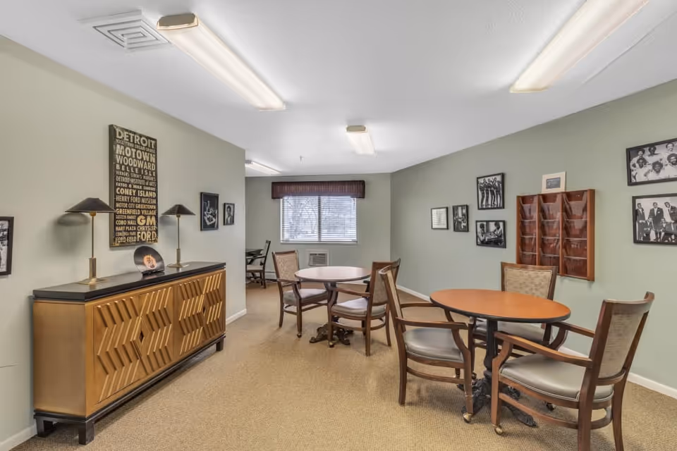 Communal dining/activity room with round tables and chairs, a decorative sideboard, and framed photos on muted green walls.