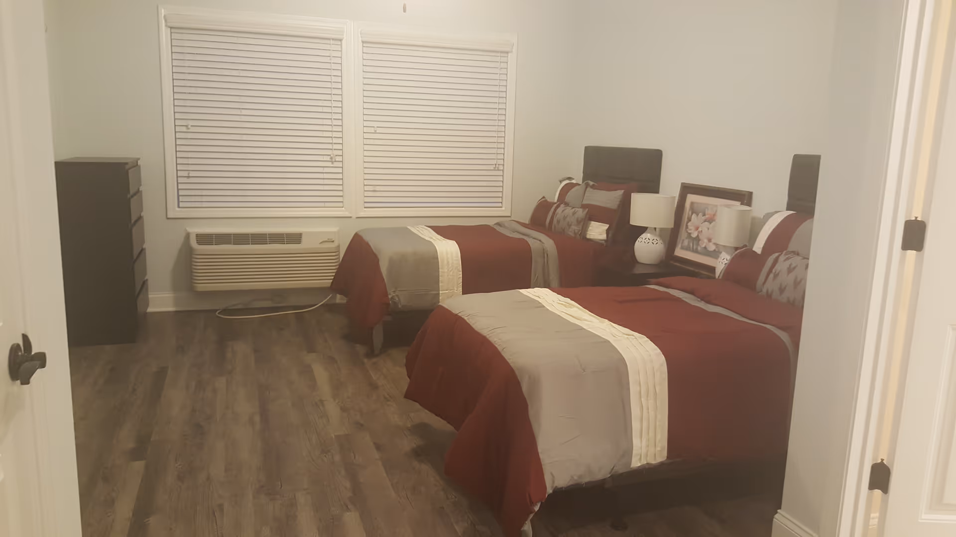 A bedroom with two twin beds featuring matching red and gray bedding. Between the beds is a nightstand with a white lamp and a framed floral picture resting against the wall. There is a window with closed blinds on the far wall, a black dresser to the left, and a wooden floor.