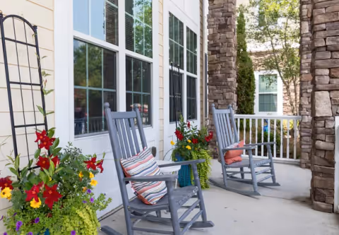 A porch area with two gray rocking chairs, each with a colorful striped cushion. There are vibrant flower pots with red, yellow, and purple flowers placed near the chairs. The porch is adjacent to a building with large windows and stone pillars, surrounded by greenery and trees.