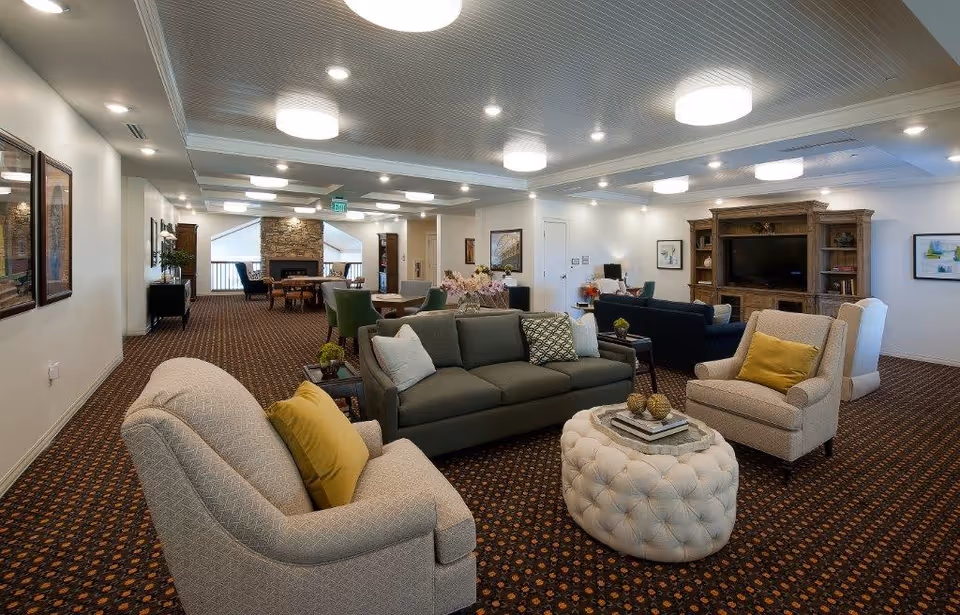 A spacious and well-lit senior living common area with multiple seating arrangements including sofas and armchairs with cushions. There is a large wooden entertainment center with a TV, a round tufted ottoman in the center, and framed artwork on the walls. The room has a patterned carpet and ceiling lights, with a stone fireplace visible in the background near a dining table and chairs.