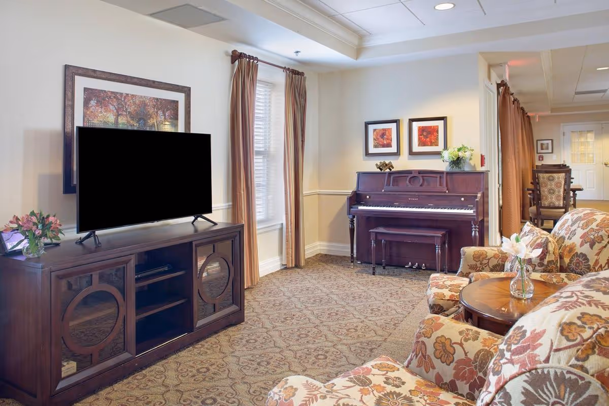 A cozy living room area in a senior living facility featuring a floral patterned sofa and armchair, a wooden coffee table with a small vase of flowers, a dark wood piano with a matching bench, two framed floral artworks on the wall above the piano, a large flat-screen TV on a wooden TV stand, and windows with beige curtains allowing natural light into the room.