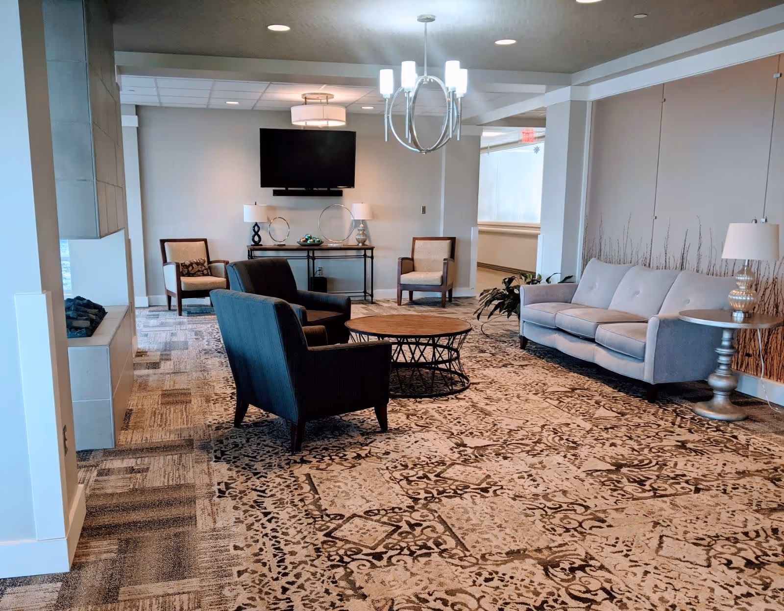 A spacious living room area in a senior living facility with a patterned carpet, a gray sofa, two dark armchairs, two beige armchairs, a round wooden coffee table, side tables with lamps, a wall-mounted flat screen TV, and modern ceiling light fixtures.