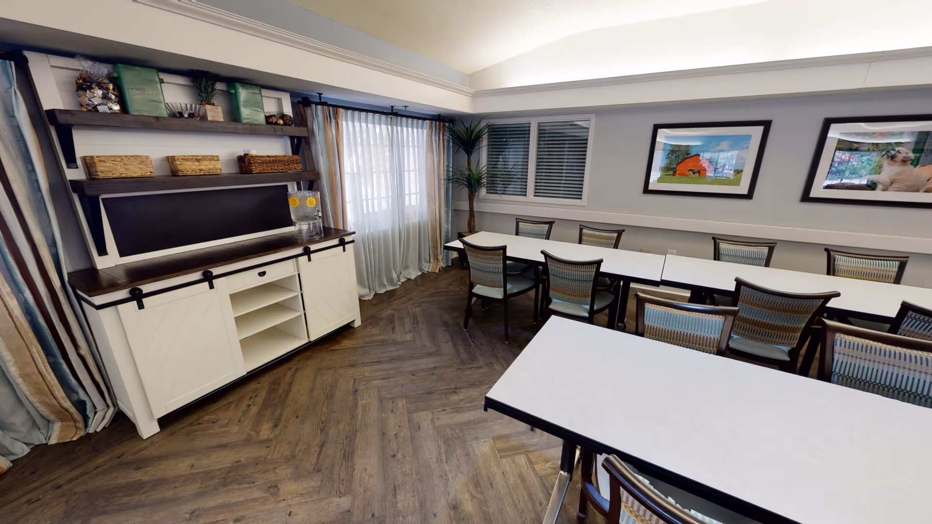 Bright communal dining room with long tables and chairs, a buffet cabinet with open shelving, framed artwork, and large windows with curtains.