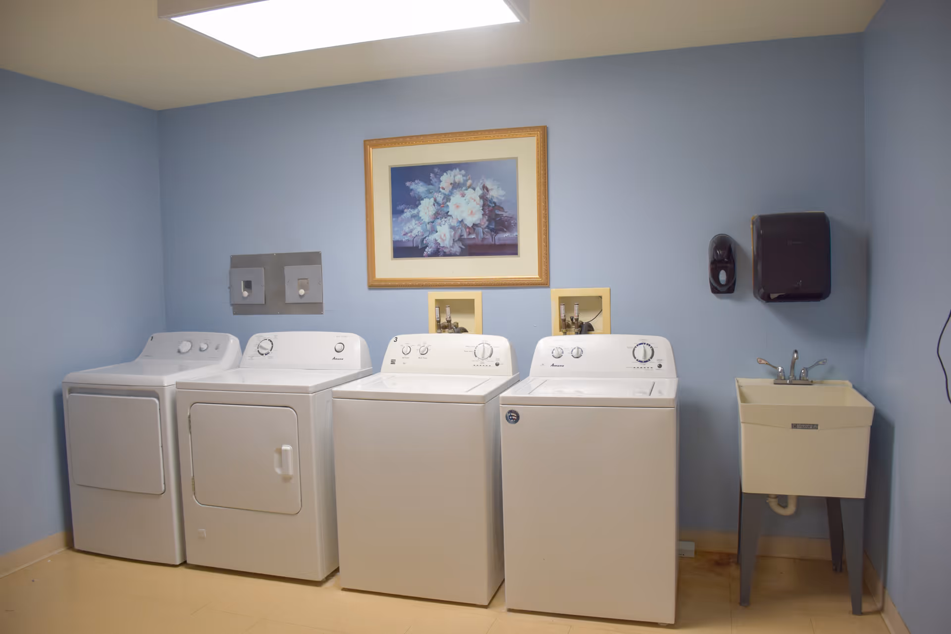 A laundry room with four white washing machines and dryers lined up against a light blue wall. Above the machines is a framed floral painting. To the right, there is a utility sink with a faucet, a soap dispenser, and a paper towel dispenser mounted on the wall.