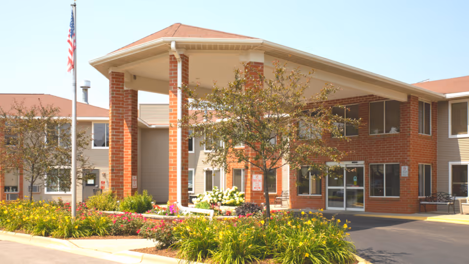 Front exterior view of Layton Terrace Senior Living facility showing a two-story building with brick and beige siding, a covered entrance supported by brick columns, landscaped flower beds, a flagpole with an American flag, and a paved driveway.