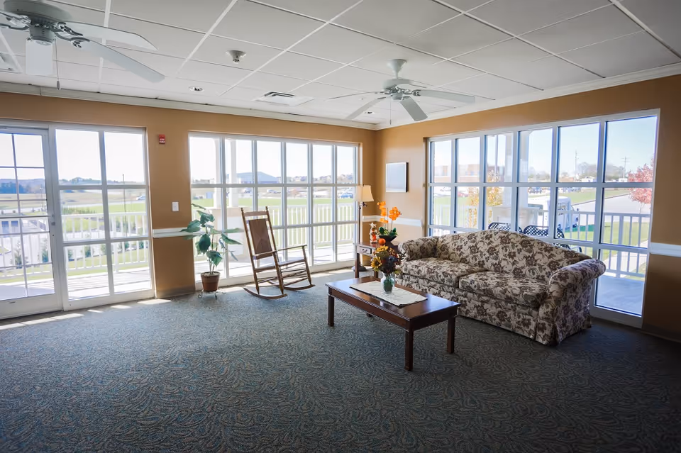 Sunlit common living area with a floral sofa, rocking chair, coffee table, and large windows overlooking the grounds.