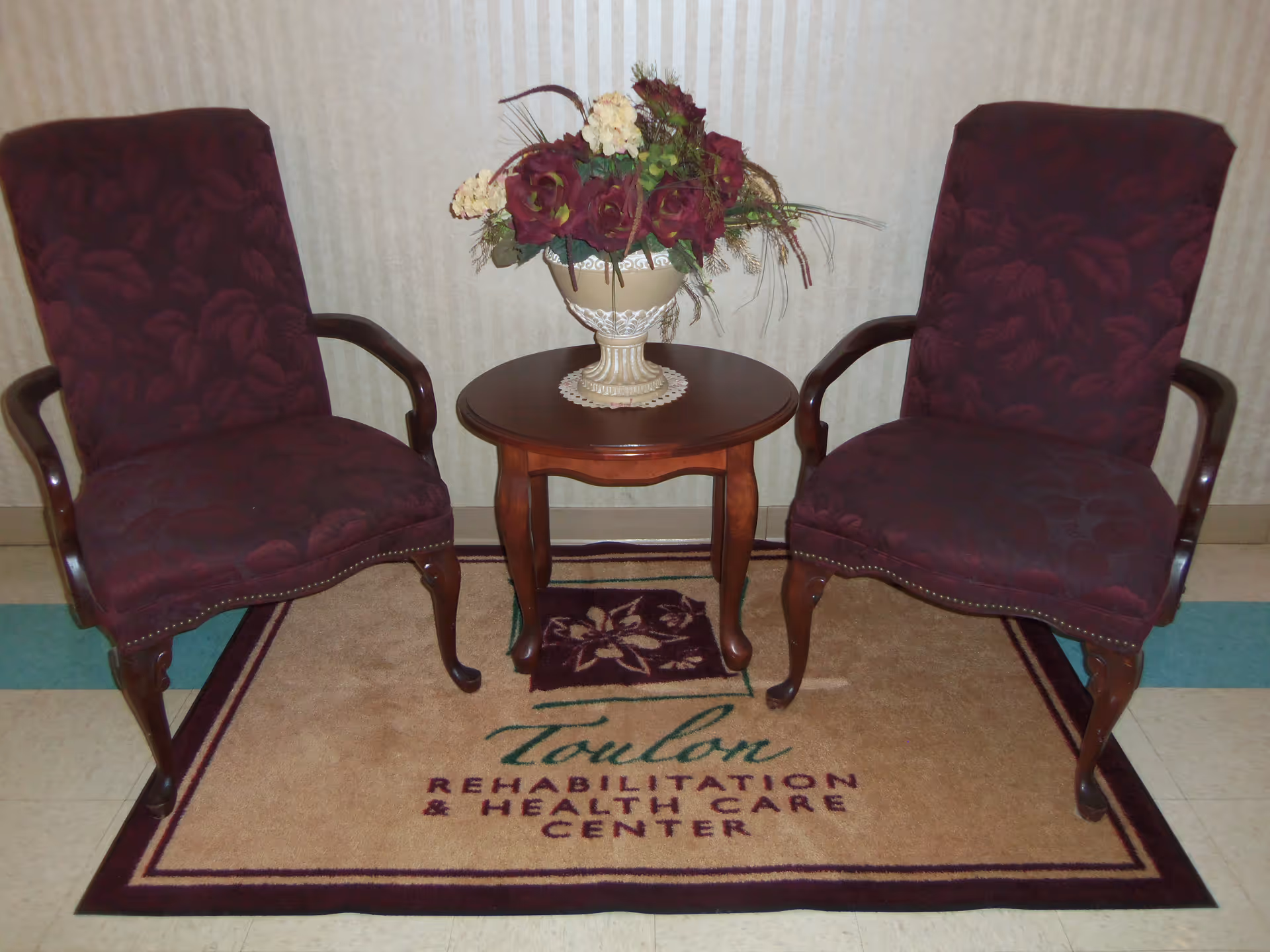 Two upholstered armchairs with dark red floral patterns are positioned on either side of a small wooden round table with a decorative flower arrangement in a vase. The furniture is placed on a beige rug with the text 'Toulon Rehabilitation & Health Care Center' written on it. The background features a light-colored striped wallpaper.