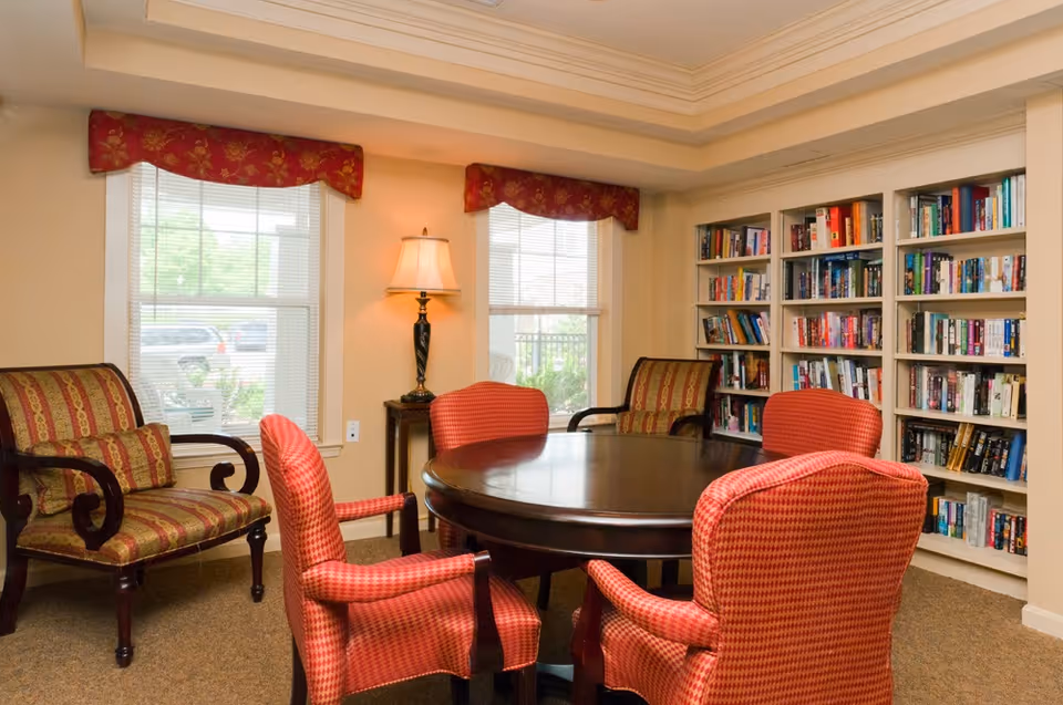 A cozy reading or meeting room with a round wooden table surrounded by four red upholstered chairs. Two additional patterned armchairs are placed near two windows with red valances. A floor lamp stands between the windows, and a large built-in bookshelf filled with books lines one wall.