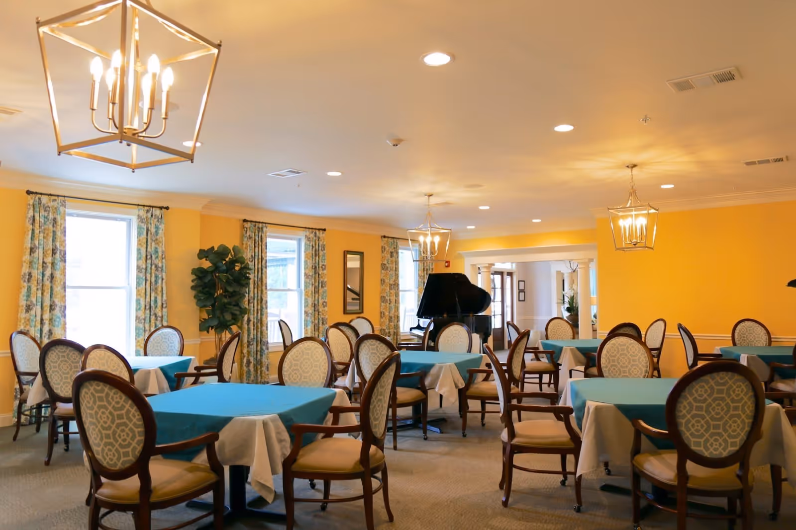 A bright dining room with yellow walls and multiple tables covered with white and teal tablecloths. Each table is surrounded by wooden chairs with patterned upholstery. The room features large windows with floral curtains, elegant hanging light fixtures, and a black grand piano in the background.