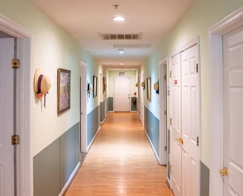 A well-lit hallway in an elder care facility with light green and white walls, wooden flooring, several closed white doors on both sides, framed pictures and hats hanging on the walls, and an exit door at the far end.