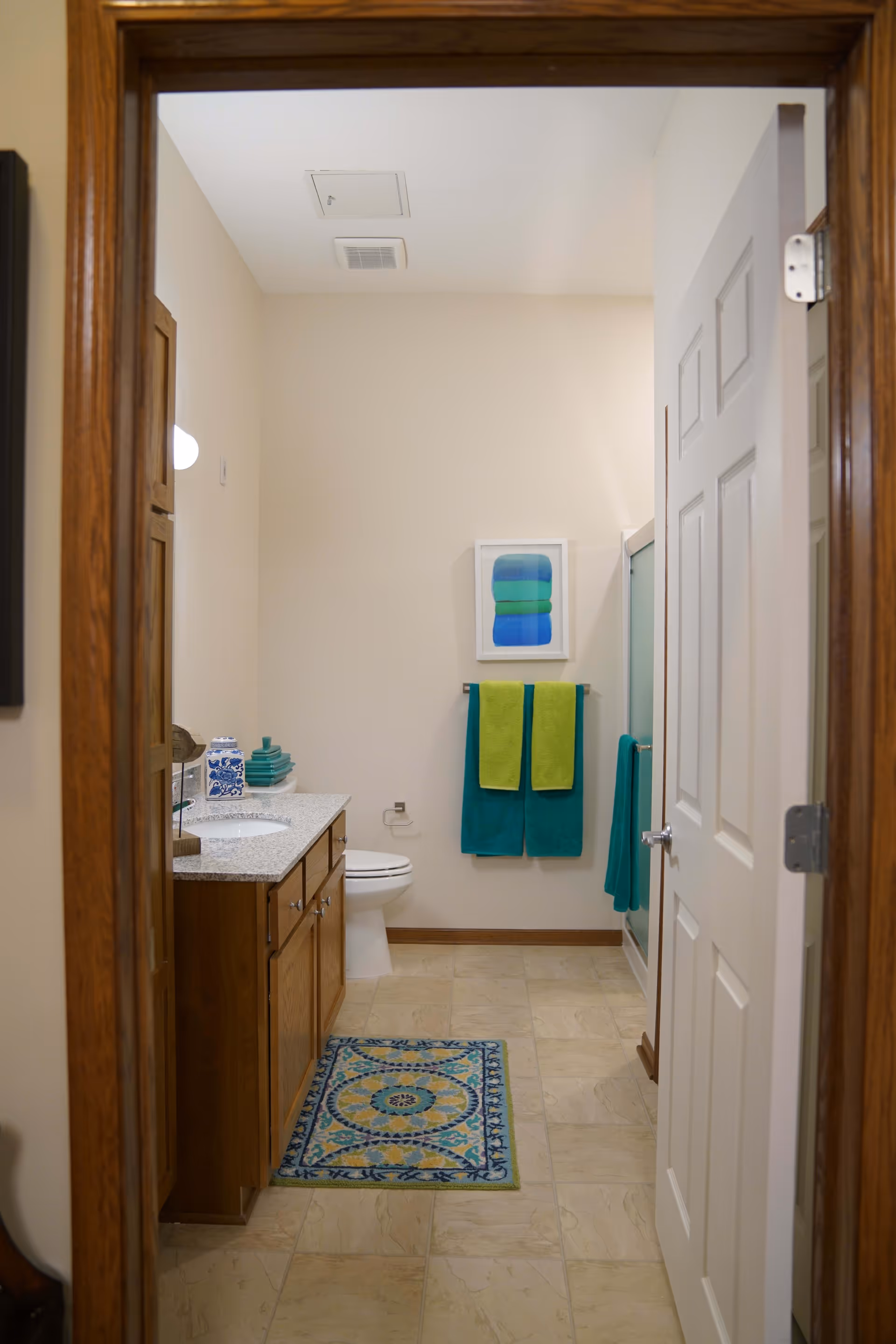 View into a bathroom with beige tiled floor, wooden cabinetry with a granite countertop, a toilet, and a glass-enclosed shower. Two green and two teal towels hang on the wall, with a framed abstract artwork above them. A decorative blue and yellow rug is on the floor in front of the sink.