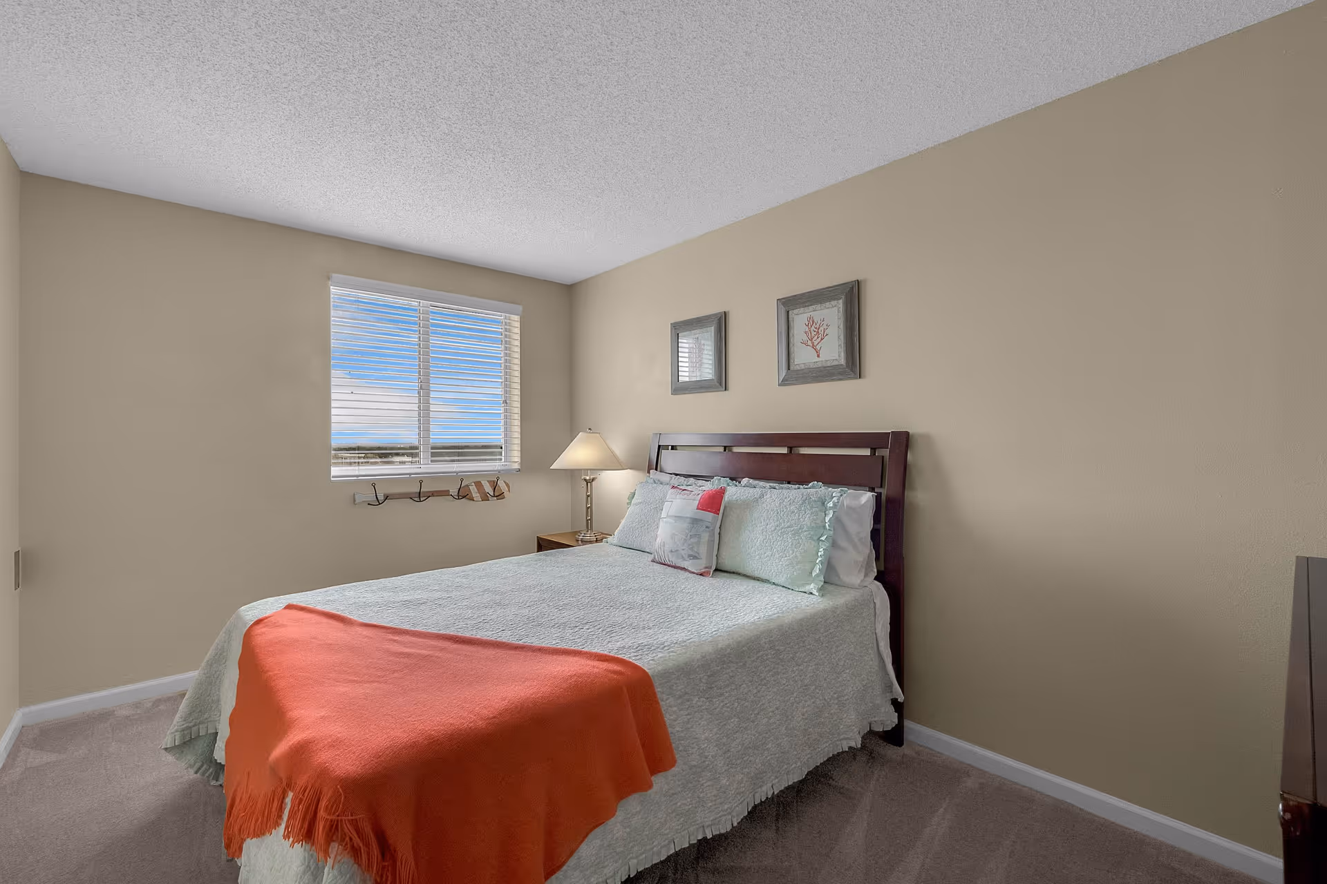 A bedroom with a neatly made bed featuring a light blue quilt and an orange throw blanket at the foot. There are two framed pictures above the wooden headboard. A window with white blinds lets in natural light, and a bedside table with a lamp is positioned next to the bed. The walls are painted beige and the floor is carpeted.