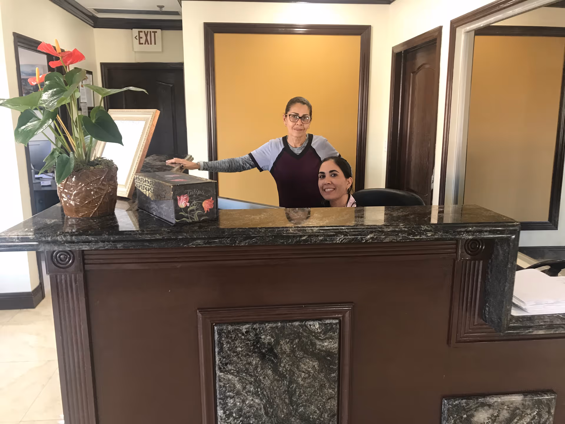 Two women behind a dark marble reception desk in an office setting. One woman is standing and the other is seated, both smiling. There is a potted plant and a decorative box on the desk. The background features a yellow wall panel, dark wooden door frames, and an exit sign above a door.