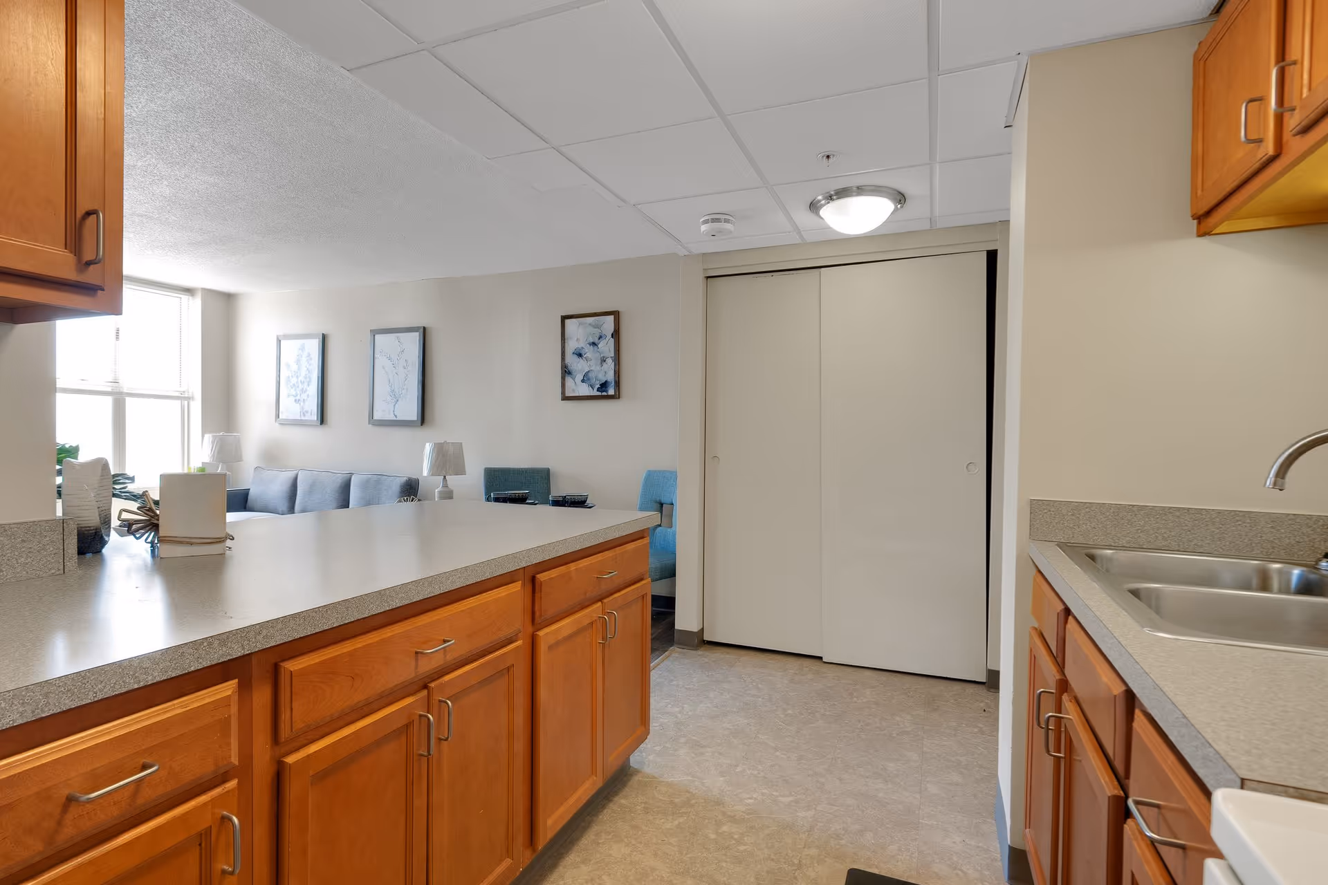 View of a kitchen area with wooden cabinets and a countertop extending into a living room with a blue couch, two framed pictures on the wall, and two blue chairs. The kitchen has a double sink and a window letting in natural light.