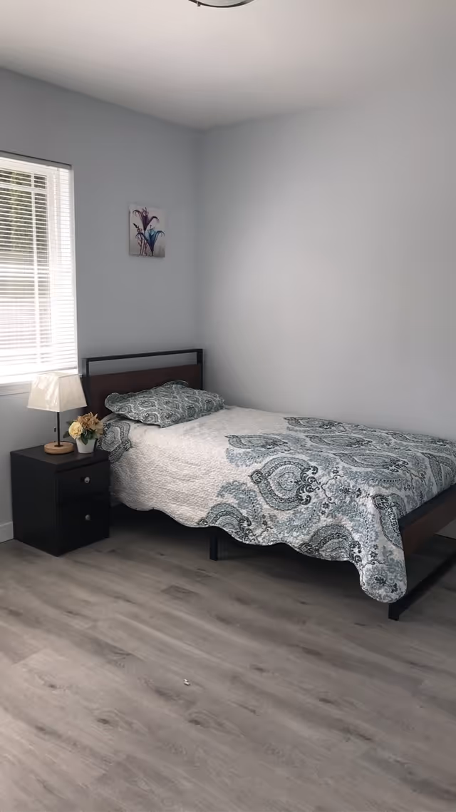 A simple bedroom with a single bed covered in a white and blue patterned quilt. Next to the bed is a black nightstand with two drawers, a small lamp, and a small flower pot. A window with white blinds is on the left wall, and a small floral painting hangs above the bed on the light gray wall. The floor is light wood.