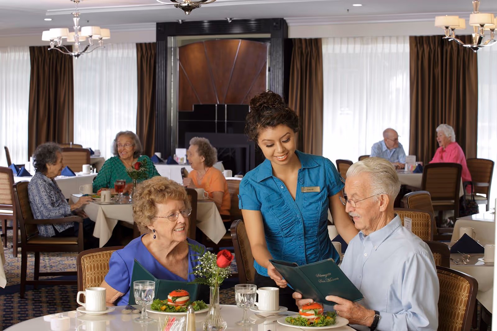 A dining room in a senior living facility where an elderly man and woman are seated at a table with plates of food. A smiling waitress in a blue uniform is showing the man a menu. Other elderly residents are seated at tables in the background, engaged in conversation. The room has large windows with white curtains and brown drapes, chandeliers, and a fireplace.