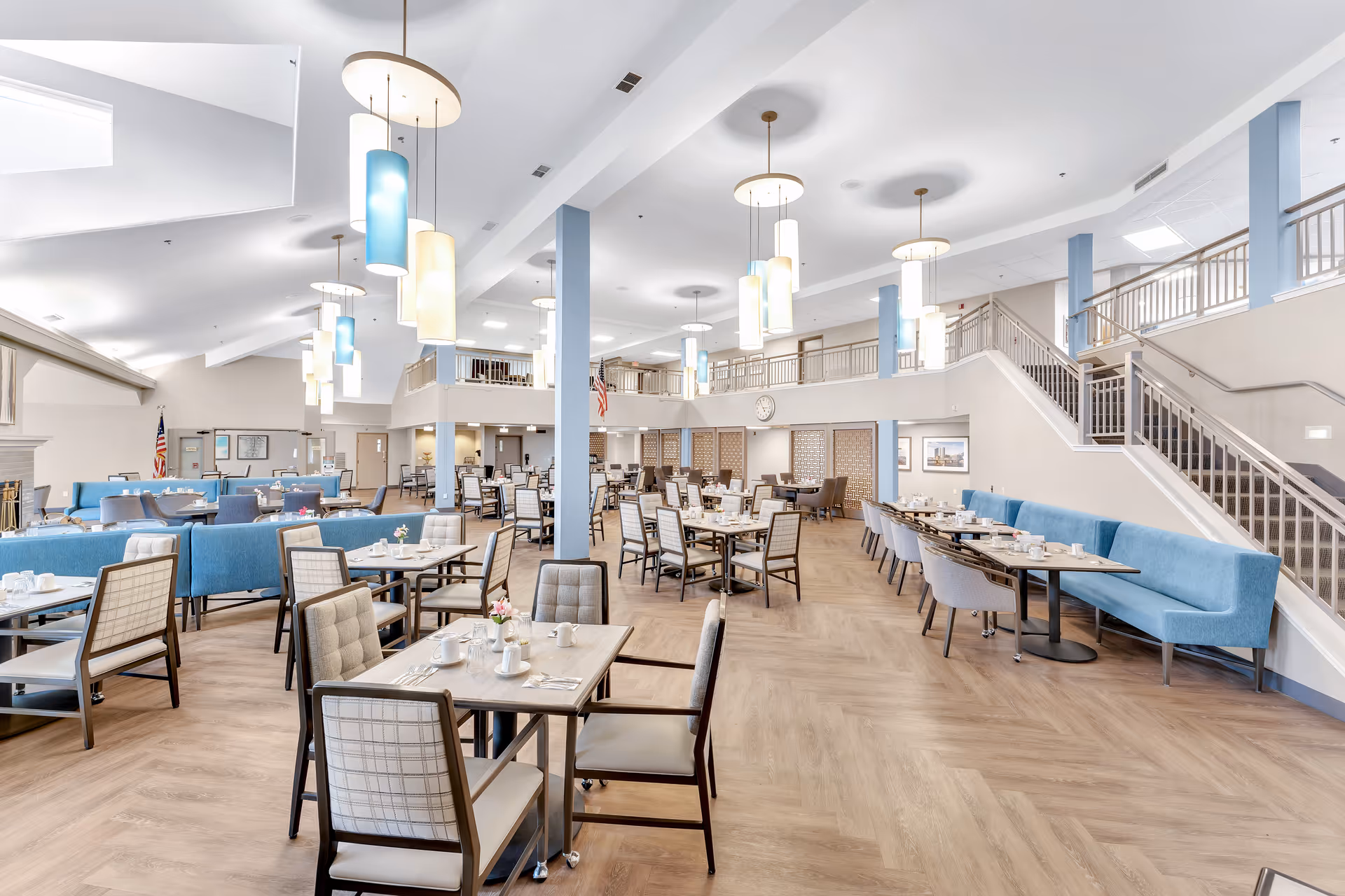 A spacious and bright dining room in a senior living facility with multiple tables and chairs arranged neatly. The room features modern pendant lighting, blue cushioned bench seating along the walls, and a staircase leading to an upper level. The floor has a light wood pattern, and the walls are painted in soft neutral tones. There are American flags and framed pictures on the walls.