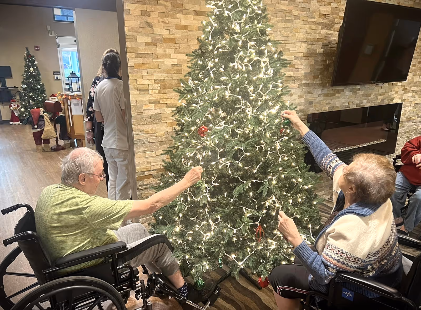 Two elderly individuals in wheelchairs decorating a large, lit Christmas tree in a cozy indoor setting with a stone accent wall and a mounted TV. In the background, there is another decorated Christmas tree and holiday decorations, with two staff members standing nearby.