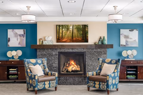 A cozy living room area with a lit fireplace in the center, flanked by two patterned armchairs with cushions. Above the fireplace is a large framed picture of a sunlit forest. On either side of the fireplace are wooden cabinets with decorative items and artwork hanging on teal-colored walls.