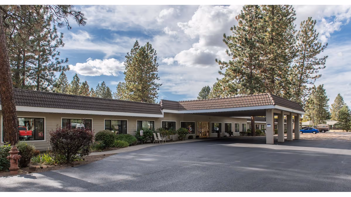 Exterior view of a single-story building with a covered entrance and several windows, surrounded by tall pine trees and a paved driveway under a partly cloudy sky.