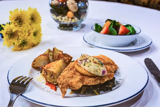 A plated meal consisting of breaded fried chicken topped with a stuffed potato half, roasted potato wedges, and a side salad with watermelon and greens in a separate bowl on a white tablecloth. A fork and knife are placed beside the plate, and a small bouquet of yellow flowers and a decorative glass bowl with assorted items are in the background.