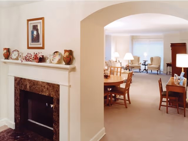 Interior view of a senior living facility showing a cozy living room and dining area. The foreground features a fireplace with decorative vases and plates on the mantel and a framed picture above it. Through an archway, there is a round wooden dining table with chairs, a desk with a lamp and framed photos, and a seating area with armchairs and lamps near a large window with vertical blinds.