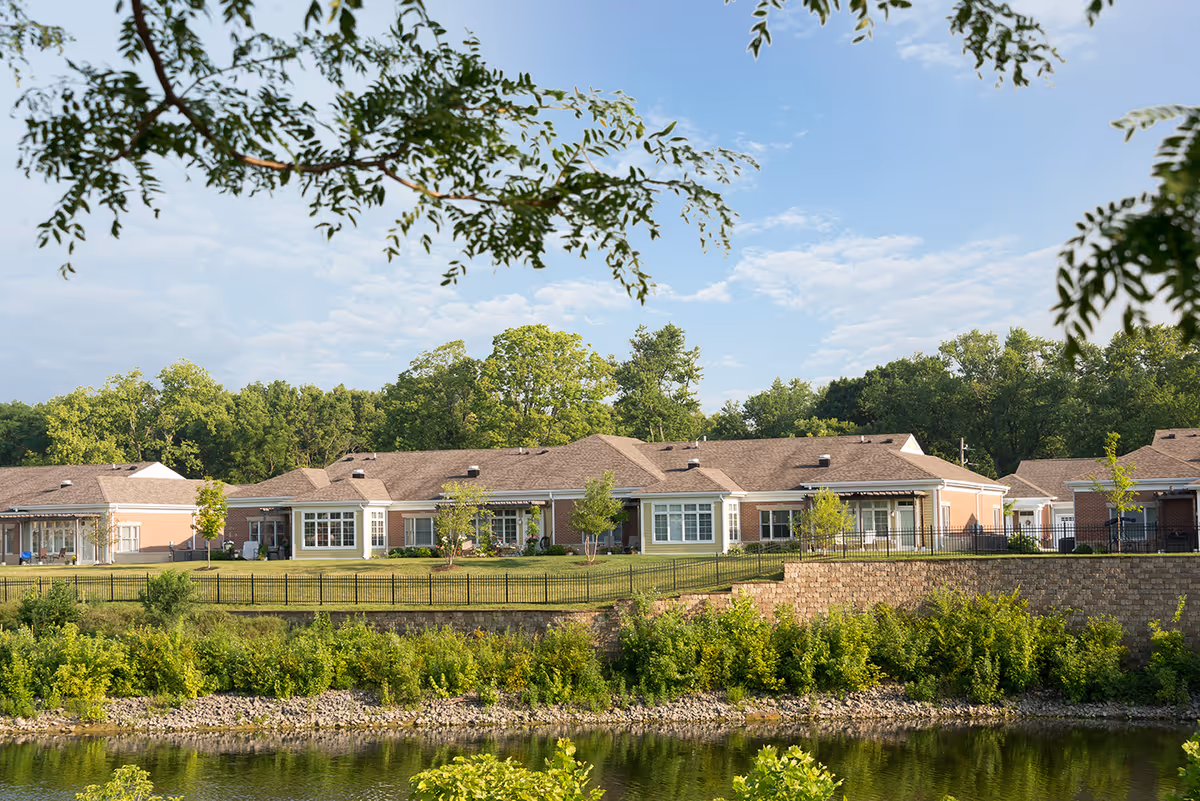 View of a single-story senior living facility building with brick exterior and multiple windows, surrounded by green trees and landscaping, with a small body of water in the foreground and a partly cloudy sky above.