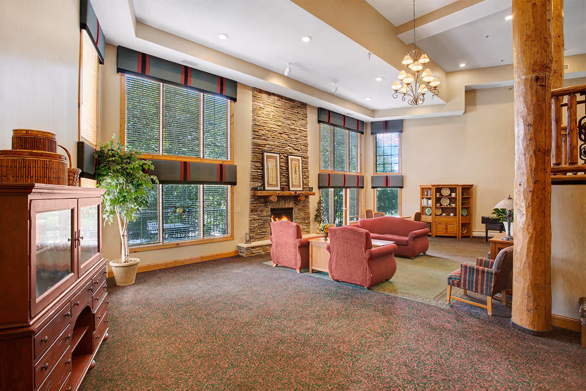 A spacious living room area with large windows covered by blinds and valances, a stone fireplace with framed artwork above it, and comfortable seating including a red sofa and armchairs around a wooden coffee table. The room features wooden furniture, a potted plant, a chandelier, and a cabinet with decorative plates. The carpet is patterned and the walls are light-colored.