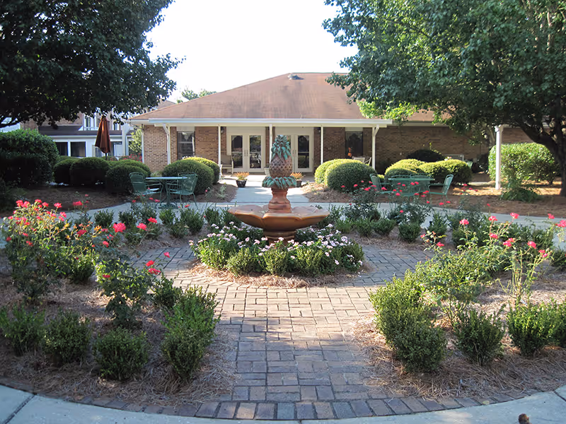A landscaped outdoor garden area with a central brick-paved circular path surrounding a decorative pineapple-shaped fountain. The garden is bordered with green bushes and flowering plants with pink and red blooms. In the background, there is a single-story brick building with a covered porch and several glass doors and windows. Green metal chairs and tables are placed near the building under the shade of trees.