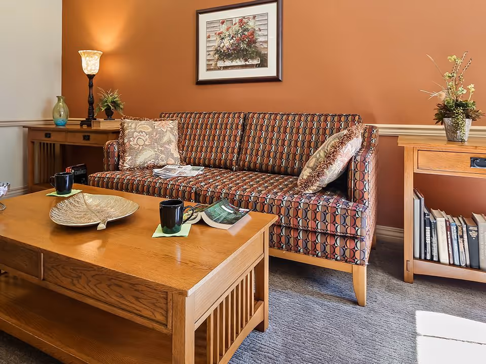 A cozy living room with a patterned sofa, wooden coffee table holding mugs and decor, side tables, lamp, and framed artwork on an orange wall.
