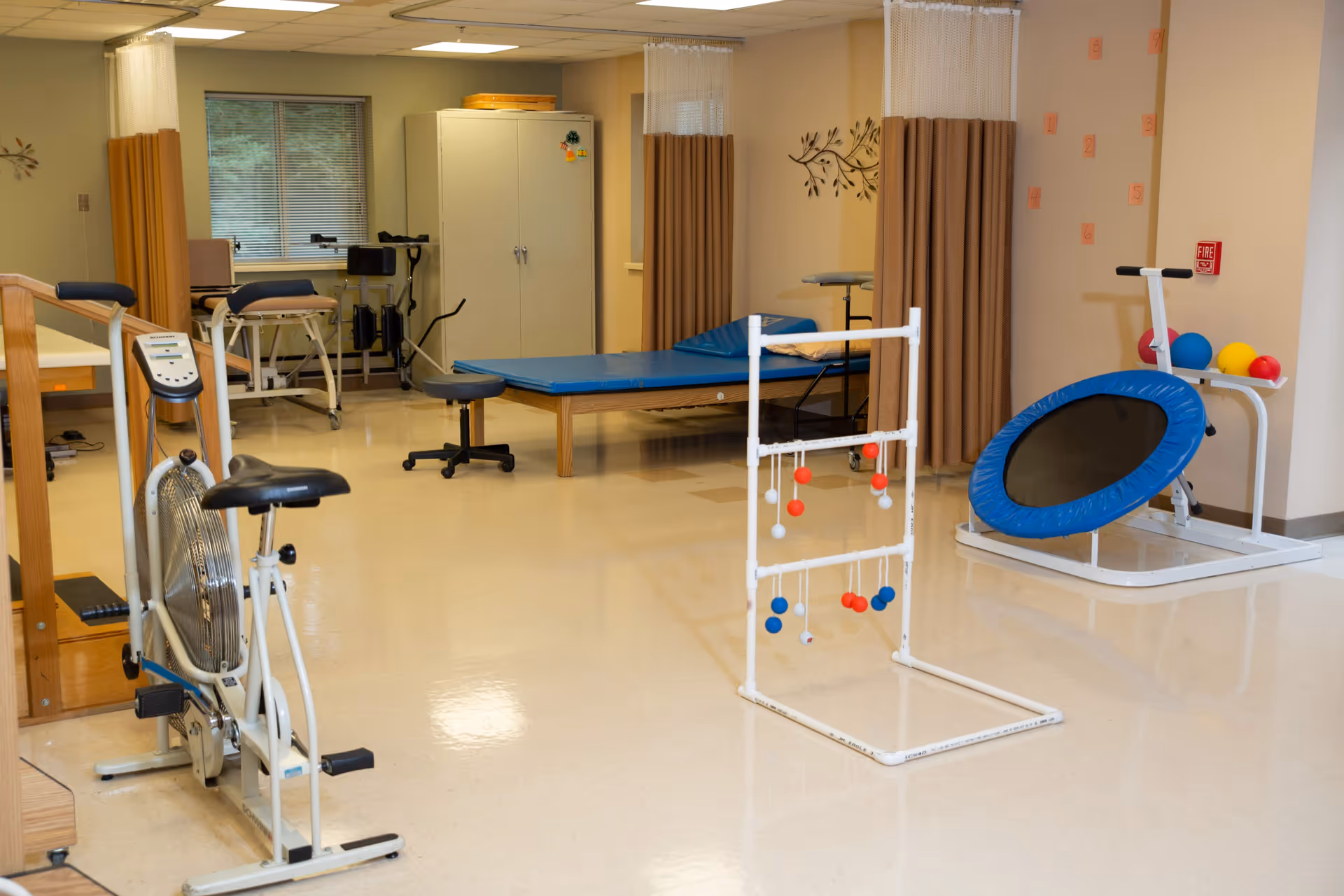 A physical therapy or rehabilitation room with exercise equipment including a stationary bike, a small trampoline, a ladder toss game, therapy tables, stools, and privacy curtains. The room has beige walls and a window with blinds.