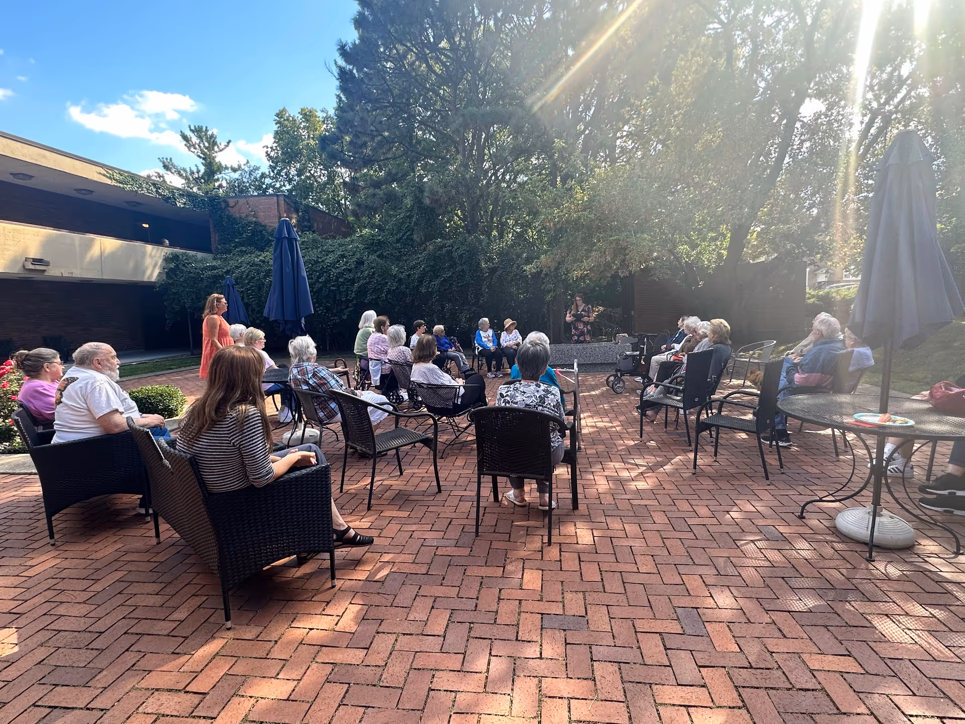 A group of elderly people seated in a semi-circle on a brick patio outdoors, listening to a woman standing and speaking. The area is surrounded by trees and greenery, with patio tables and umbrellas nearby under a sunny sky.