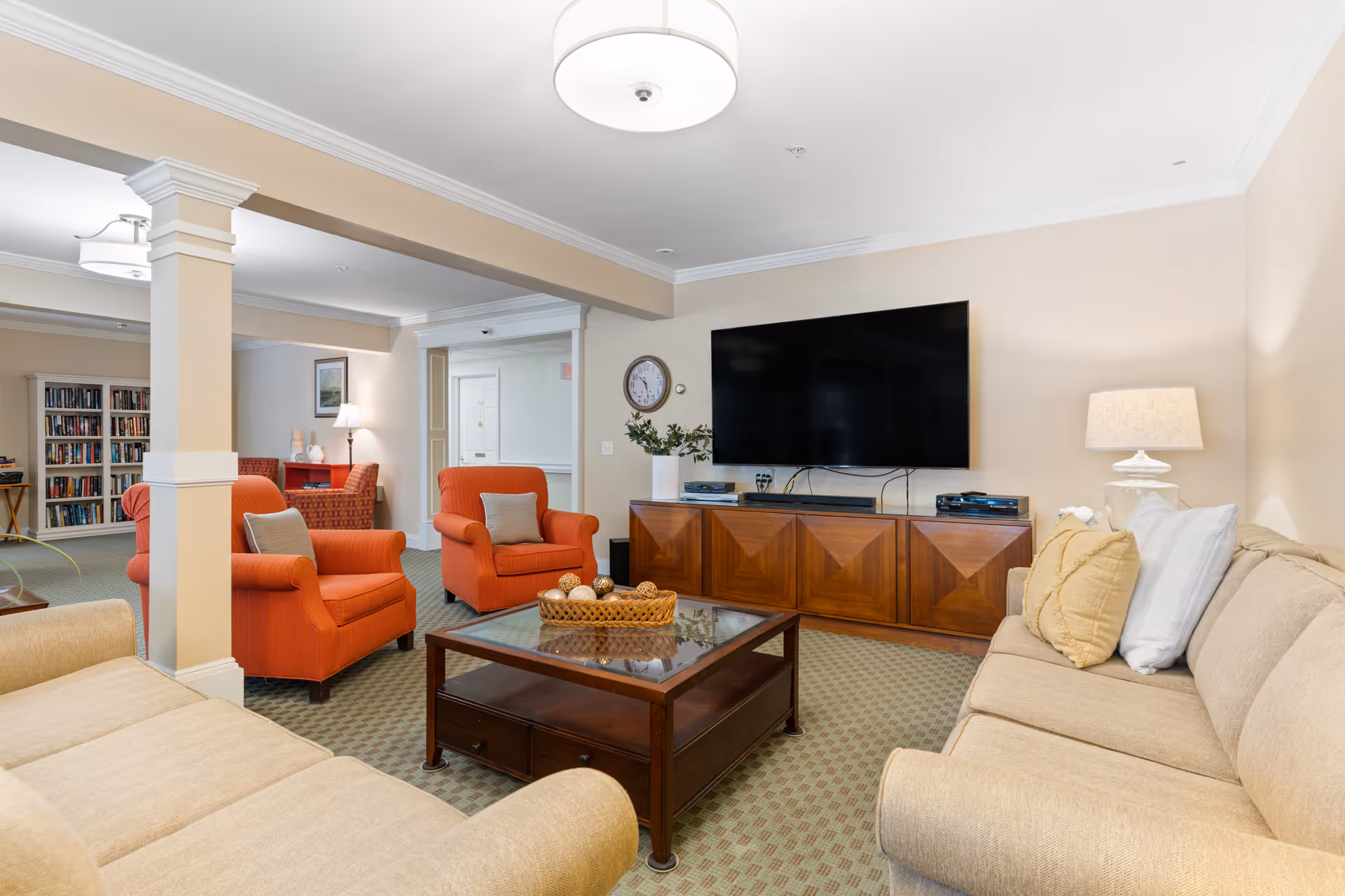 Bright communal living room with beige sofas, orange armchairs, a wall-mounted TV on a wooden console, and a central coffee table.