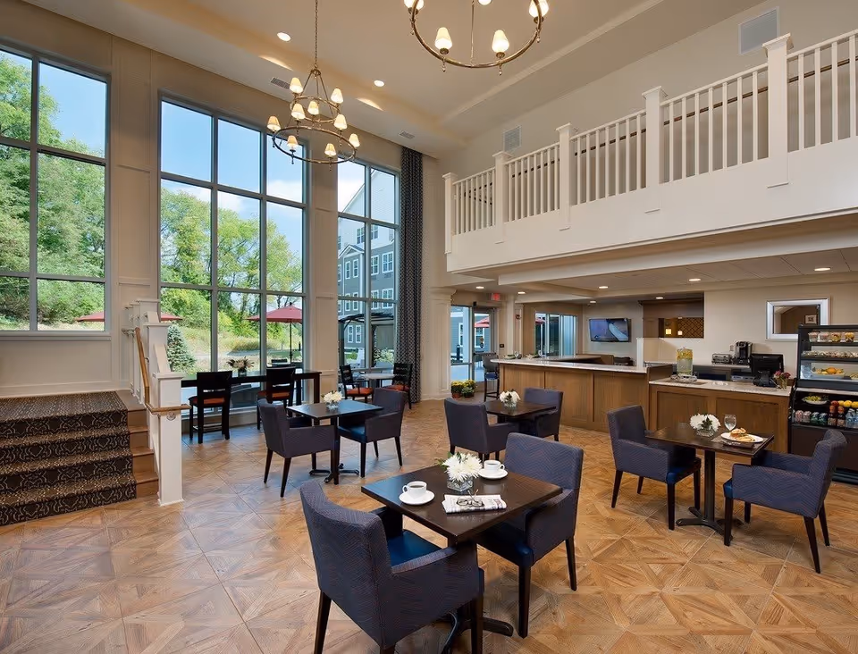 A bright and spacious dining area with large floor-to-ceiling windows showing green trees outside. The room features several dark wood tables with blue upholstered chairs, each table decorated with a small white flower arrangement. There is a staircase with patterned carpet on the left, and a counter area with a display case of food items on the right. Two chandeliers hang from the ceiling, and there is a balcony railing above the counter area.