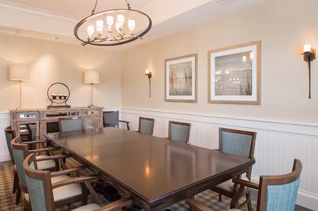A well-lit dining room featuring a long dark wood table surrounded by upholstered chairs, a sideboard, framed artwork, and a chandelier.