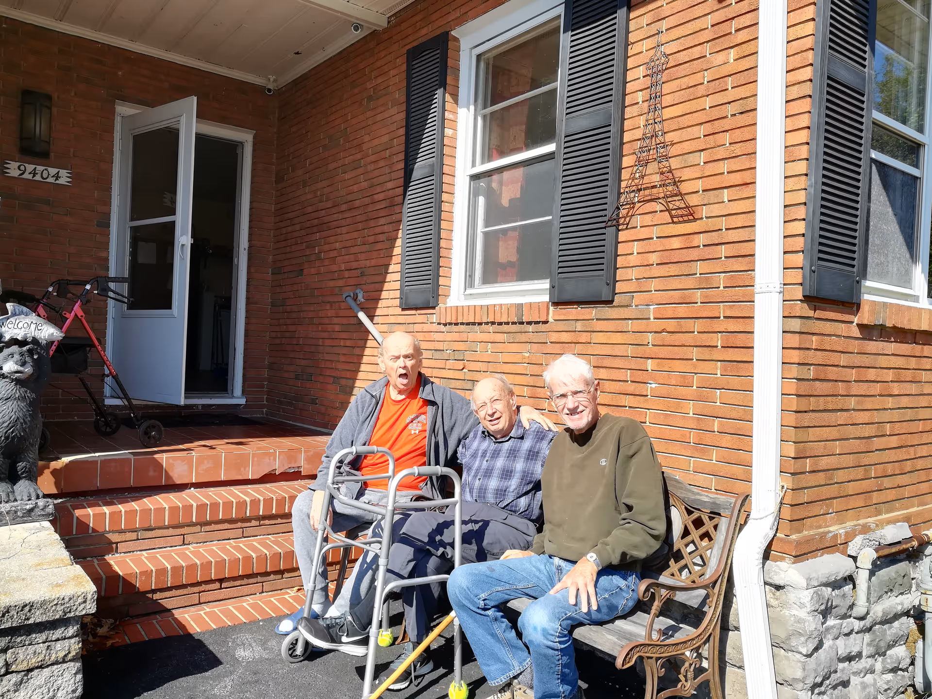 Three elderly men sitting by a walker on the brick front steps of a house.
