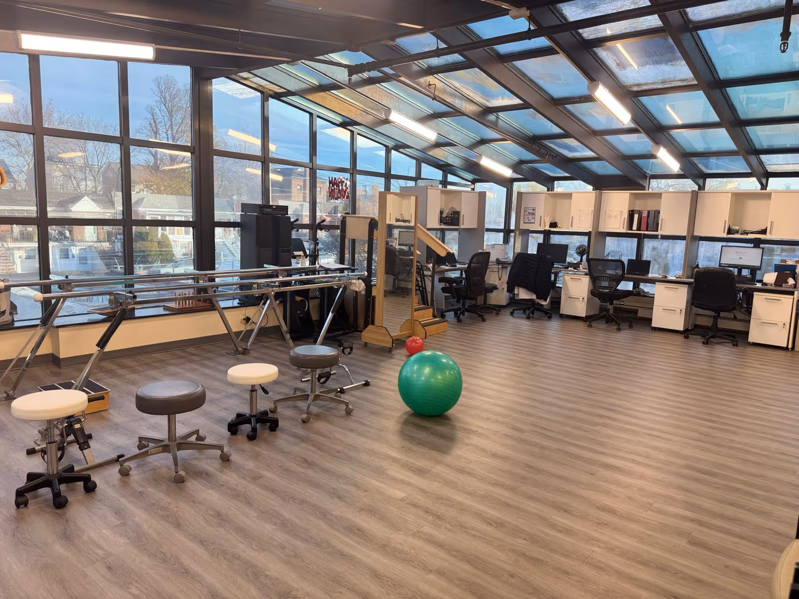 A spacious rehabilitation room with large windows and a glass ceiling allowing natural light to fill the space. The room features parallel bars for walking exercises, several stools on wheels, a green exercise ball, and a red ball. In the background, there are multiple desks with office chairs and computer monitors, as well as shelves with various items.