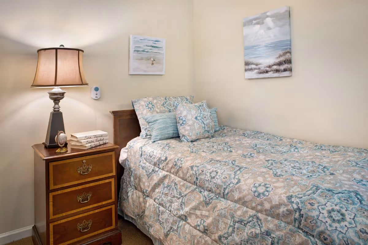 A cozy bedroom with a single bed covered in a patterned bedspread and matching pillows. Next to the bed is a wooden nightstand with three drawers, a table lamp, a small clock, and a stack of books. Two paintings depicting beach scenes hang on the light-colored walls.