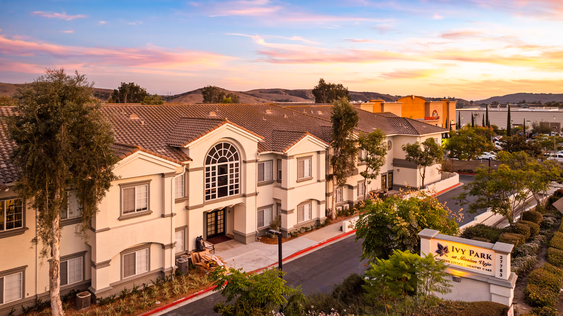 Exterior view of Ivy Park at Mission Viejo senior living facility during sunset, showing a two-story building with a tiled roof, large arched window above the entrance, surrounding trees, and a sign with the facility's name and address.