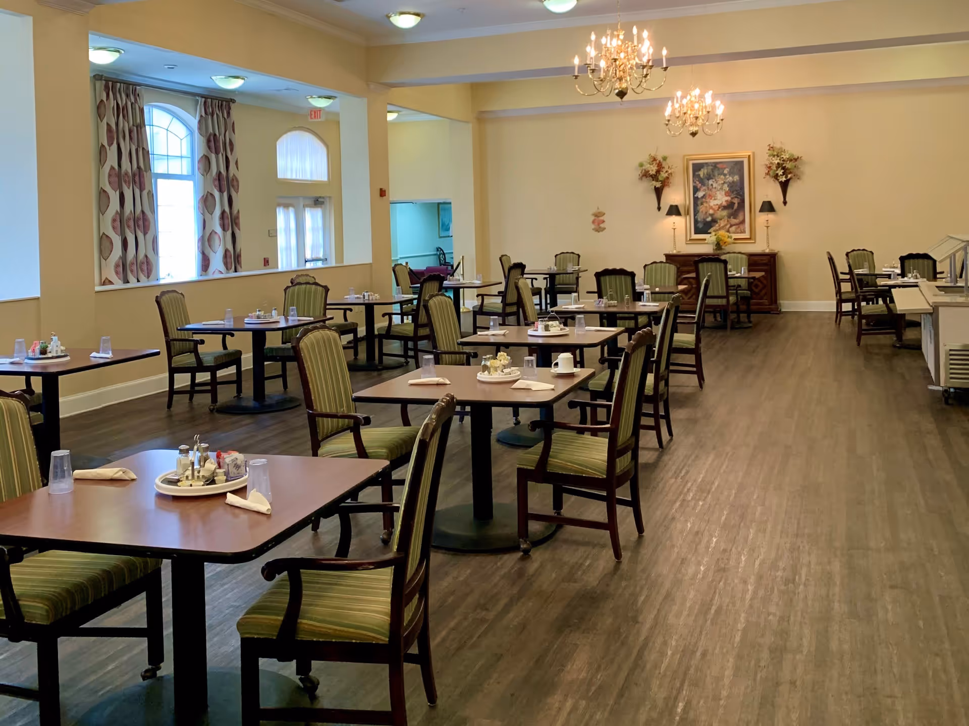A spacious dining room with multiple square tables, each set with cups, napkins, and condiments. The room has wooden flooring, green cushioned chairs, chandeliers hanging from the ceiling, and floral decorations on the far wall. Large windows with patterned curtains allow natural light into the room.