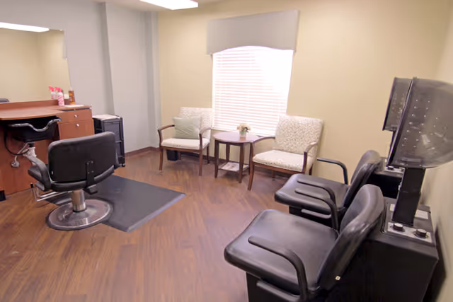 Interior view of a salon area in a senior living facility with a black salon chair in front of a wooden counter with hair products, two black hair dryer chairs, and a small seating area with two cushioned chairs and a round table near a window with blinds.