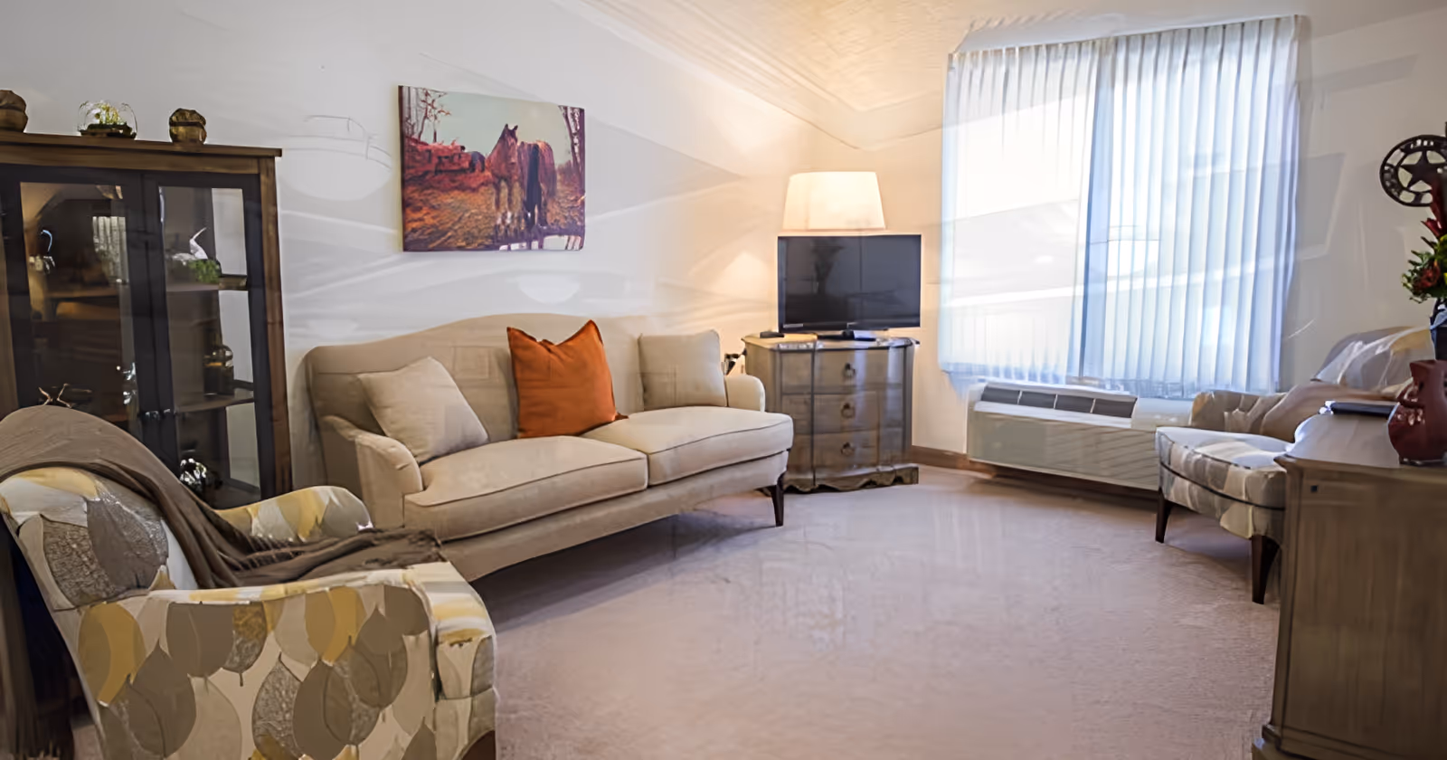 A bright sitting room with a sofa, patterned armchairs, a TV on a small dresser, display cabinet and a large window with sheer curtains.
