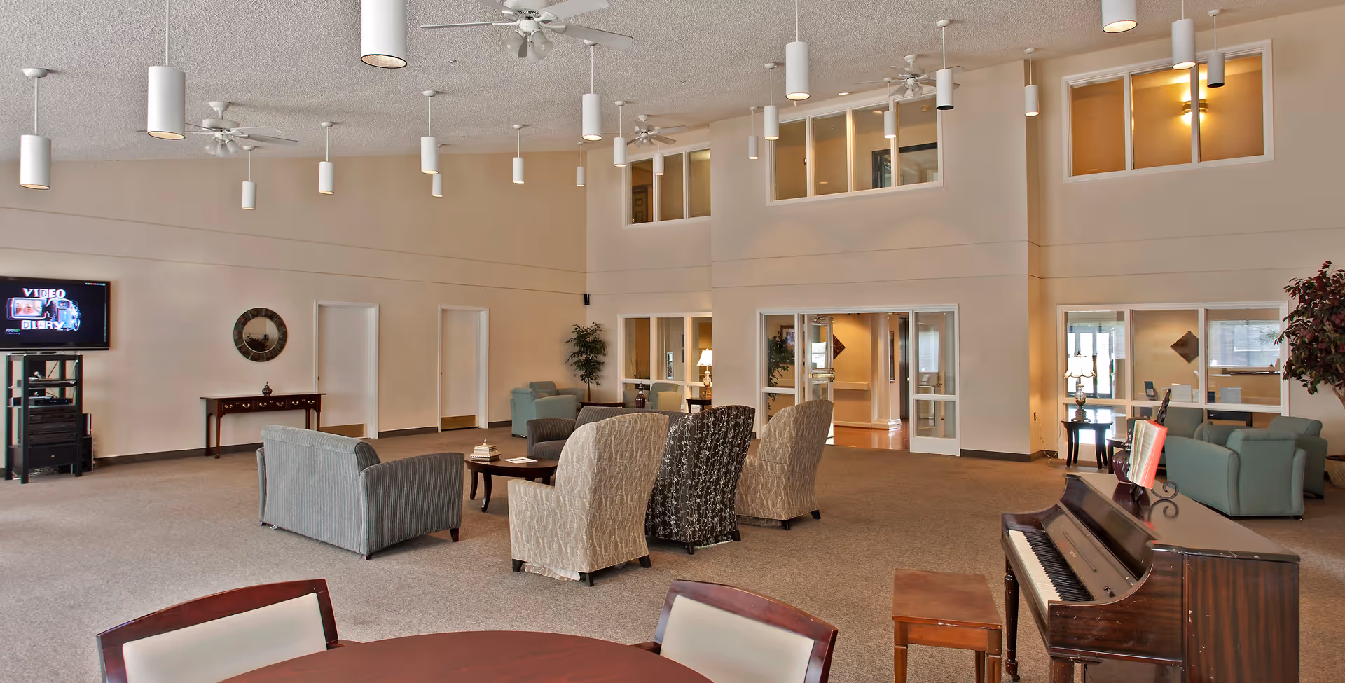 A spacious senior living facility common area with multiple seating arrangements including armchairs and sofas, a piano with a bench, a round table with chairs in the foreground, ceiling fans and pendant lights, and a TV mounted on the wall displaying 'Video Diary'. The walls are beige with large windows and doors leading to other rooms.