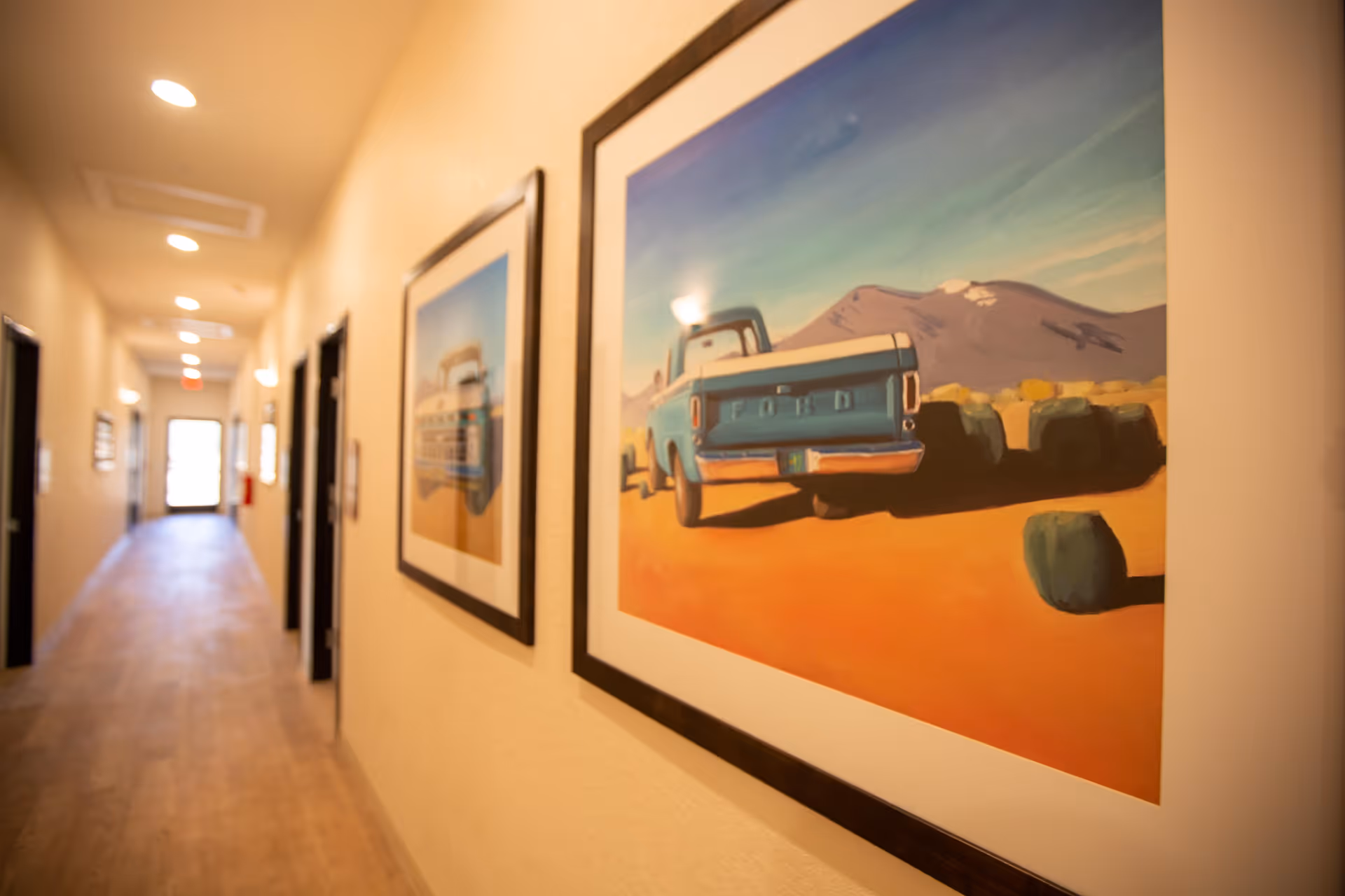 A well-lit hallway in a senior living facility with wooden flooring and beige walls. The hallway features several doors on both sides and framed paintings of vintage trucks and rural landscapes hanging on the right wall. The corridor extends towards a bright exit door at the far end.