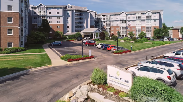 Exterior view of Freedom Village senior living community showing a multi-story residential building with a parking lot in front, landscaped greenery, and a sign that reads 'Freedom Village A Senior Living Community'.
