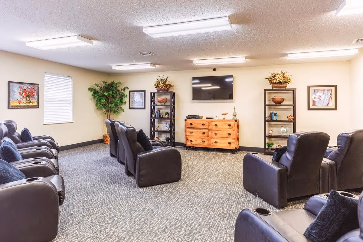 A senior living facility common room with multiple black leather recliners arranged in rows facing a wall-mounted flat screen TV. The room has beige walls, carpeted floor, two black shelving units with decorative items, a wooden dresser, framed artwork, and a potted plant near a window with blinds.