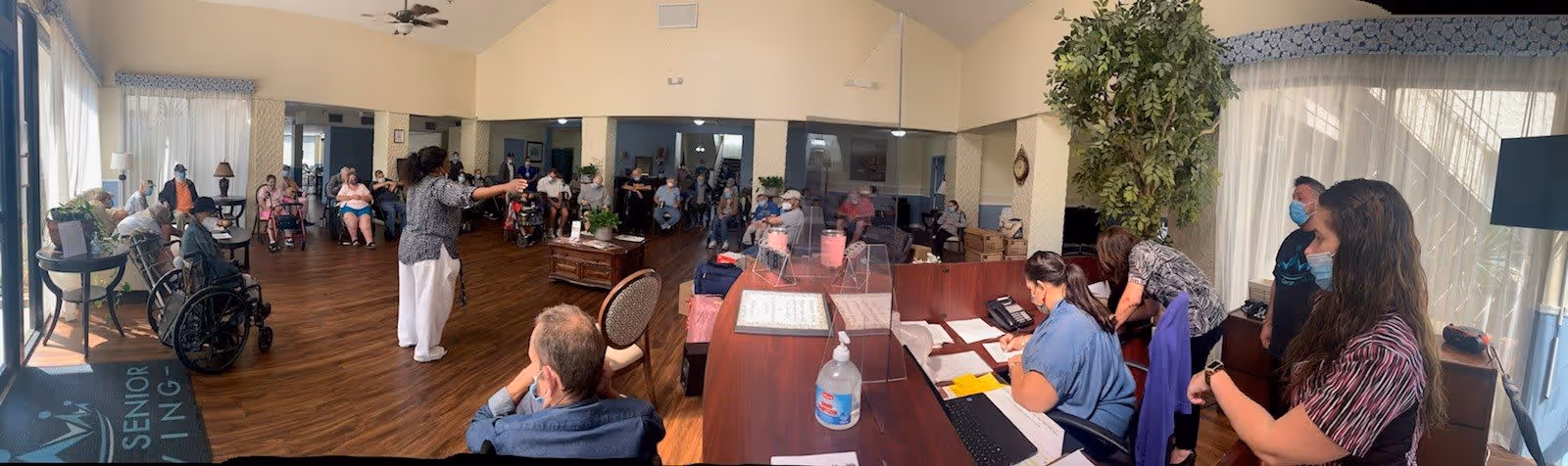 A large room in a senior living facility with many elderly residents seated in chairs and wheelchairs arranged in a semi-circle. A woman stands in the center addressing the group. On the right side, staff members are seated and standing behind a reception desk with office supplies and a hand sanitizer bottle. The room has large windows with curtains, wood flooring, and a ceiling fan. There is a large potted plant near the reception area.