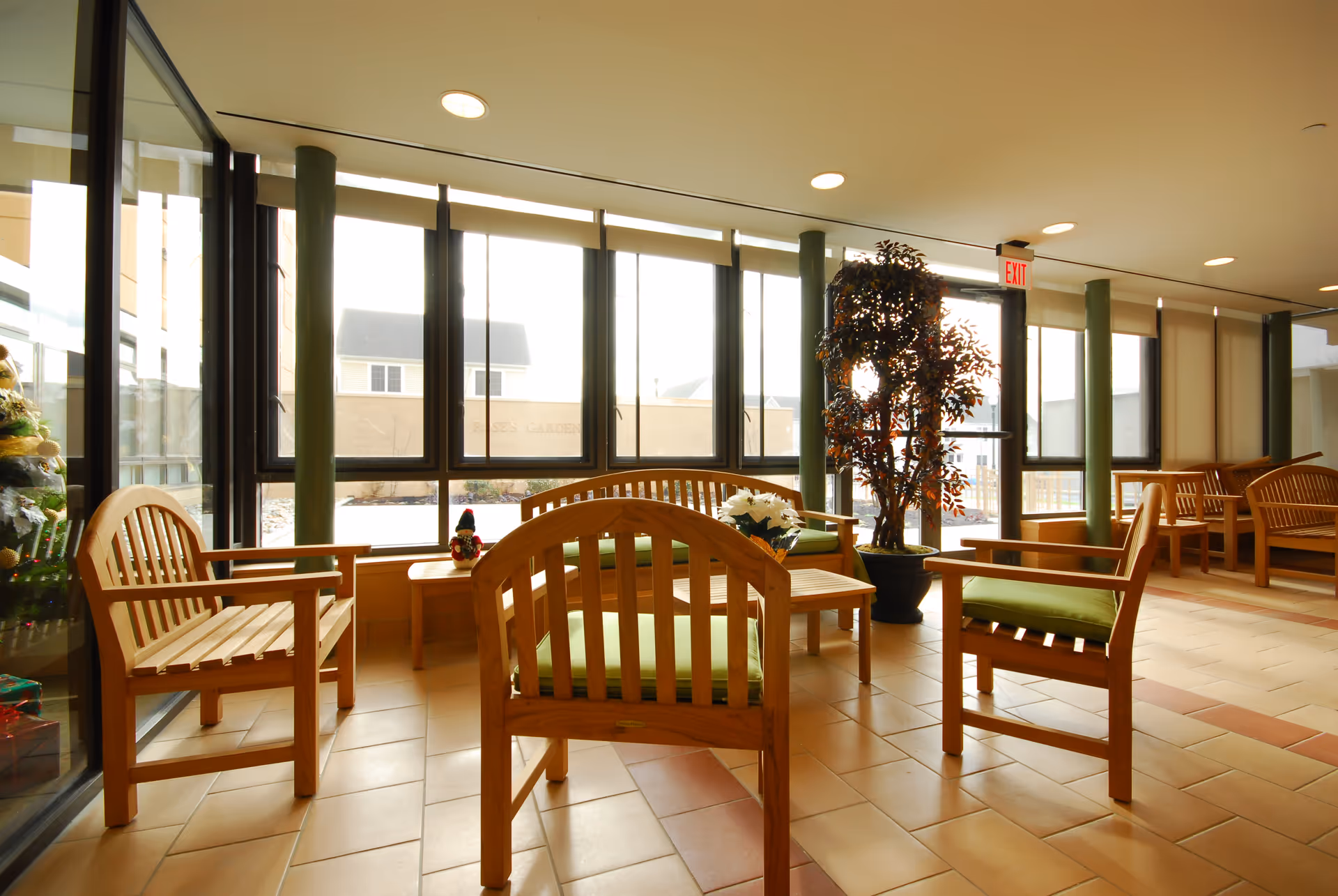 A bright indoor seating area with wooden chairs and benches featuring green cushions, a small wooden table with a flower pot, large windows letting in natural light, and a potted plant near an exit door.